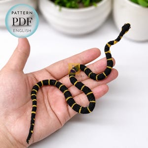Könnte beinhalten: Eine kleine gehäkelte Schlange mit einem sich wiederholenden Muster aus schwarzen und gelben horizontalen Streifen. Die Schlange ist in einer Schlangenform gewunden. Ein kreisförmiges blaues Abzeichen in der oberen linken Ecke enthält den Text PATTERN PDF ENGLISH.