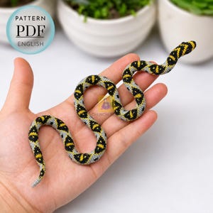 Puede incluir: Un juguete de serpiente de ganchillo con un patrón repetitivo de diamantes negros y amarillos sobre un fondo gris claro. La serpiente está enroscada en forma de S. Una insignia azul circular en la esquina contiene el texto PATTERN PDF ENGLISH.