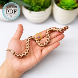 Puede incluir: Un juguete de serpiente de ganchillo con un patrón repetitivo de manchas marrones sobre un fondo color crema. La serpiente tiene un cuerpo delgado y sinuoso y ojos pequeños y oscuros. Una insignia azul muestra el texto PATTERN PDF ENGLISH.