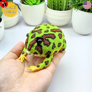 Puede incluir: Patrón de crochet para una rana verde y marrón con ojos negros. La rana está sentada en una mano.