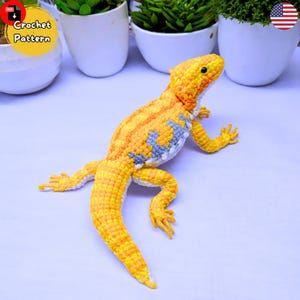 Peut inclure: Un lézard crocheté aux tons jaune et orange, avec des touches de bleu et de blanc. Le reptile fait main a des pattes détaillées, une longue queue et est posé sur une surface claire. L'image comprend le texte "Crochet Pattern".