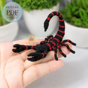 May include: A small, crocheted scorpion figurine featuring a black body with vibrant red stripes along its tail, legs, and pincers. The text PATTERN PDF ENGLISH is visible in the upper left corner, and the logo CHUNCHUNDRA is in the centre.
