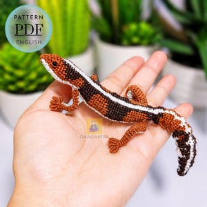 Puede incluir: Un lagarto amigurumi de ganchillo con un cuerpo estampado en marrón, marrón oscuro y blanco, una cola larga y cuatro patas pequeñas. El artículo es un patrón PDF en inglés, como indica el texto circular en la esquina superior izquierda.
