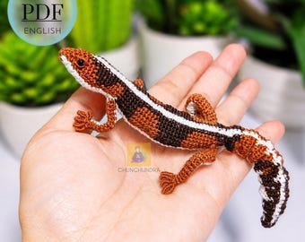 Könnte beinhalten: Ein gehäkeltes Eidechsen-Amigurumi mit einem braunen, dunkelbraunen und weißen Musterkörper, einem langen Schwanz und vier kleinen Beinen. Der Artikel ist eine PDF-Anleitung auf Englisch, wie durch den kreisförmigen Text oben links angezeigt.