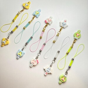 May include: A set of nine colourful phone charms featuring various animal and floral designs. Each charm is attached to a braided cord with a gold bead accent.