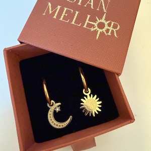 Puede incluir: Pendientes de aro dorados con colgantes celestiales, incluyendo una luna creciente con un pequeño cristal y un sol. Presentados en una caja marrón con el texto "SIAN MELLOR".