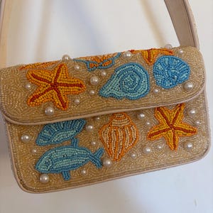 May include: A rectangular shoulder bag covered in gold-toned beads featuring a marine-themed design. The surface displays beaded motifs of orange starfish, blue fish, and various seashells, accented with scattered white faux pearls throughout.