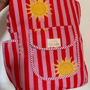 May include: A quilted backpack featuring vertical red and pink stripes with a yellow sun pattern. The front includes a pocket with a white label reading SIAN MELLOR and a red and white striped trim along the edges of the pocket and side panel.