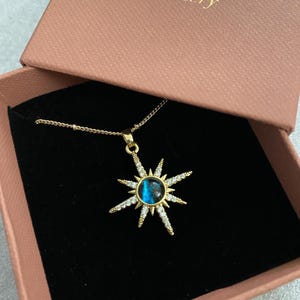 Könnte beinhalten: Eine goldene Sternburst-Halskette mit einem blauen, schillernden Stein in der Mitte. Die Halskette befindet sich in einer rosa Box mit dem Text "Sian Mellor Jewellery".