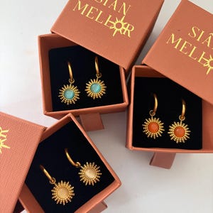 Puede incluir: Pendientes de aro dorados con colgantes de sol en varios colores, incluyendo turquesa, naranja y crema, expuestos en cajas color coral. Las cajas llevan el texto "SIÂN MELER".