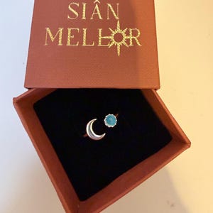 May include: A silver ring with a crescent moon design and a turquoise gemstone, presented in an open, rust-colored box with the gold text "SIÂN MELLOR". The ring sits on a black velvet interior.