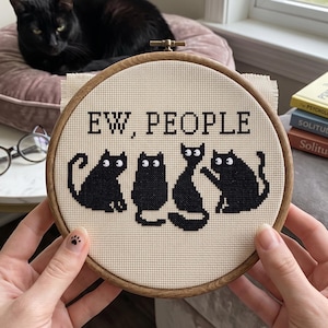 Pu&ograve; includere: Un telaio da ricamo circolare in legno contenente un pezzo di tessuto Aida color crema con un motivo a punto croce. Il design presenta il testo nero "EW, PEOPLE" sopra quattro gatti neri stilizzati con occhi bianchi su sfondo chiaro.
