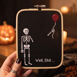 May include: A rectangular wooden embroidery hoop containing black fabric with a white cross-stitch skeleton. The skeleton is missing an arm, while a detached skeletal hand holds a red balloon. The text "Well, Shit ..." is stitched in white.