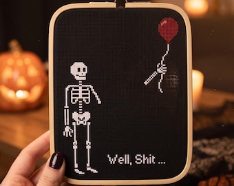以下が含まれることがあります： 黒い布地に白いクロスステッチで骸骨が刺繍された長方形の木製刺繍枠。骸骨は片腕が欠けており、切り離された骸骨の手が赤い風船を持っている。白い糸で「Well, Shit ...」という文字が刺繍されている。