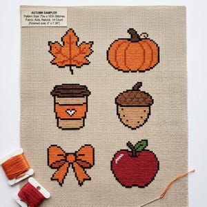 May include: An autumn sampler cross-stitch pattern on natural Aida fabric featuring an orange leaf, pumpkin, coffee cup, acorn, bow, and red apple. Text reads: AUTUMN SAMPLER, Pattern Size: 70w x 103h Stitches, Fabric: Aida, Natural, 14 Count (Finished size: 5" x 7.36").