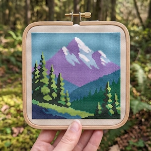 May include: A square wooden embroidery hoop containing a cross-stitch landscape. The design features a purple mountain with white snow-capped peaks, set against a light blue sky, with dark green pine trees in the foreground and a blue lake.