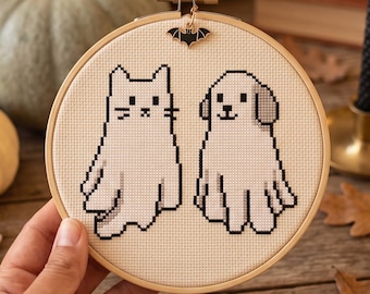 Op de afbeelding: Een ronde houten borduurring met een kruissteekontwerp van een witte kat en een witte hond, beide afgebeeld als spoken met zwarte omlijningen. Een kleine zwarte metalen vleermuisbedel is bevestigd aan de bovenkant van de spanschroef.