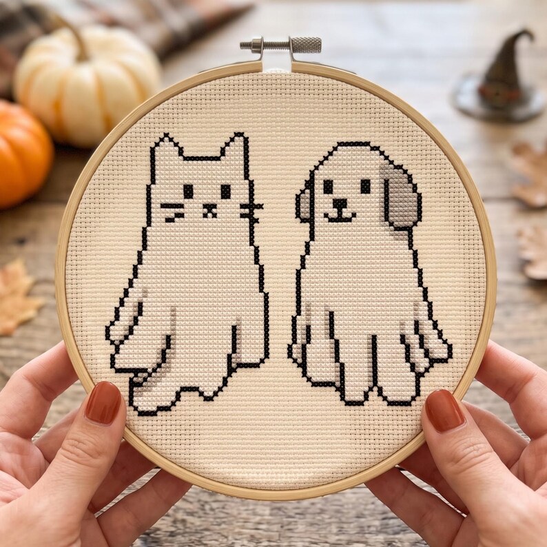 Puede incluir: Un bastidor de bordado circular de madera que contiene un patr&oacute;n de punto de cruz sobre tela de color crema. El dise&ntilde;o presenta dos figuras de estilo pixel-art con contornos negros: un gato a la izquierda y un perro con una oreja gris a la derecha.