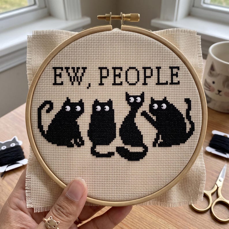 Pu&ograve; includere: Un telaio da ricamo rotondo in legno contenente un tessuto Aida beige con un disegno a punto croce. Il motivo raffigura quattro gatti neri e il testo nero "EW, PEOPLE" ricamato in uno stile di carattere pixelato nella parte superiore.