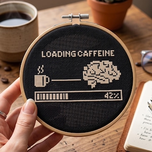 Peut inclure: Un tambour à broder circulaire en bois contenant un tissu noir avec un motif au point de croix. Le motif présente le texte LOADING CAFFEINE, une tasse de café, un cerveau et une barre de chargement indiquant 42% en fil beige clair.