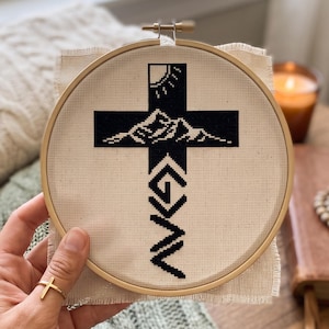 Peut inclure: Une broderie au point de croix dans un tambour en bois rond pr&eacute;sentant un motif de croix noire sur tissu beige. La croix contient un graphique de soleil et de montagnes en haut et les lettres G, N et V empil&eacute;es verticalement en dessous.