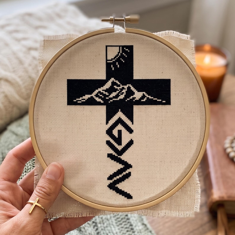 Peut inclure: Une broderie au point de croix dans un tambour en bois rond pr&eacute;sentant un motif de croix noire sur tissu beige. La croix contient un graphique de soleil et de montagnes en haut et les lettres G, N et V empil&eacute;es verticalement en dessous.