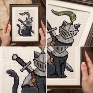 Op de afbeelding: Een ingelijst kruissteek borduurwerk met een zwarte kat in pixel-art stijl die een zilveren ridderhelm draagt met een groene en gouden pluim. De kat heeft een zwaard op de rug gebonden met een bruine leren riem op een witte achtergrond.