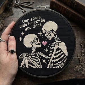 Op de afbeelding: Een ronde borduurring met zwarte stof en een kruissteekontwerp van twee skeletten die tegenover elkaar staan. Tussen hen in staat een klein roze hartje en sterretjes. De tekst luidt, Our souls didn't meet by accident.