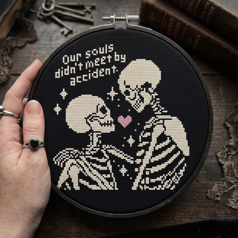 Op de afbeelding: Een ronde borduurring met zwarte stof en een kruissteekontwerp van twee skeletten die tegenover elkaar staan. Tussen hen in staat een klein roze hartje en sterretjes. De tekst luidt, Our souls didn't meet by accident.