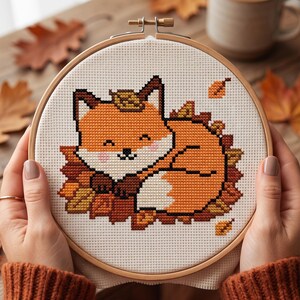 May include: A circular wooden embroidery hoop containing a cross-stitch pattern of a curled-up orange fox resting among brown and yellow autumn leaves on white fabric. The fox has a white muzzle, black eyes, and a small black nose.