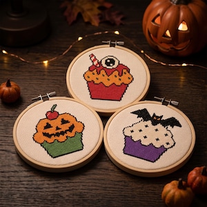 Puede incluir: Tres bastidores de bordado circulares de madera con dise&ntilde;os de punto de cruz de cupcakes con tem&aacute;tica de Halloween. Uno muestra un cupcake rojo con un globo ocular, otro un cupcake con forma de calabaza y otro un cupcake morado con un murci&eacute;lago.