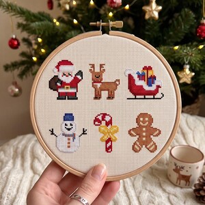 Pu&ograve; includere: Un telaio da ricamo circolare in legno contenente un motivo a punto croce su tessuto color crema. Il design presenta sei icone natalizie: Babbo Natale, una renna, una slitta rossa con regali, un pupazzo di neve, un bastoncino di zucchero e un omino di pan di zenzero.