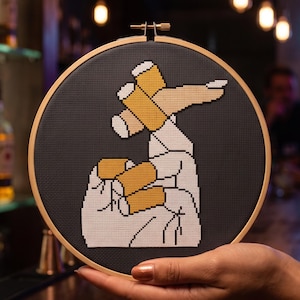 Pu&ograve; includere: Un telaio da ricamo circolare in legno contenente un disegno a punto croce su tessuto grigio scuro. Il design presenta una mano bianca stilizzata con dita color beige che regge tre oggetti cilindrici gialli, simili a bicchieri da birra.