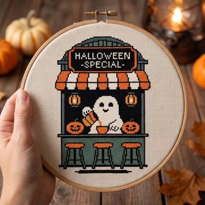 May include: A circular wooden embroidery hoop containing a cross-stitch design of a ghost serving drinks at a stand. The stand features a sign reading HALLOWEEN -SPECIAL-, orange and white striped awnings, two jack-o'-lanterns, and three stools.