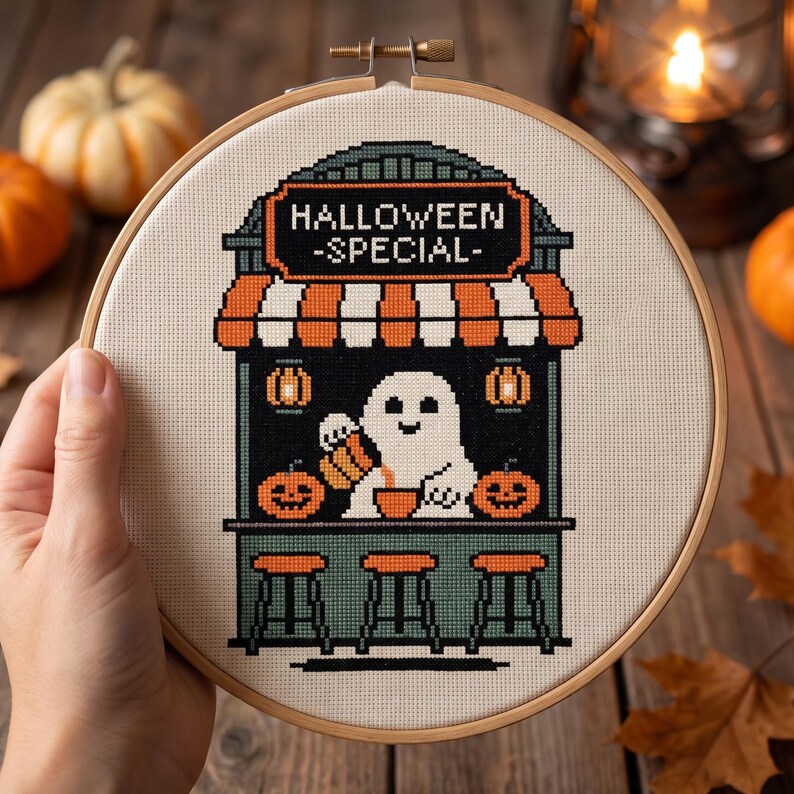 May include: A circular wooden embroidery hoop containing a cross-stitch design of a ghost serving drinks at a stand. The stand features a sign reading HALLOWEEN -SPECIAL-, orange and white striped awnings, two jack-o'-lanterns, and three stools.