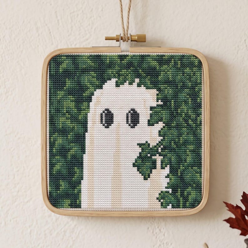 May include: A square wooden embroidery hoop containing a cross-stitch piece. The design features a white ghost figure with two dark oval eyes, positioned against a dense, textured background of various shades of green foliage and leaves.