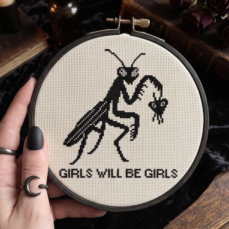 May include: A circular embroidery hoop containing a piece of off-white fabric with a black cross-stitch design of a praying mantis holding a small heart. The text "GIRLS WILL BE GIRLS" is stitched in black capital letters below the insect.