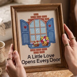 Peut inclure: Une broderie au point de croix encadr&eacute;e repr&eacute;sentant une fen&ecirc;tre avec des volets bleus ouverts, des c&oelig;urs rouges et des jardini&egrave;res sur un mur de briques. Le texte A Little Love Opens Every Door est brod&eacute; en noir sur un fond en tissu cr&egrave;me.