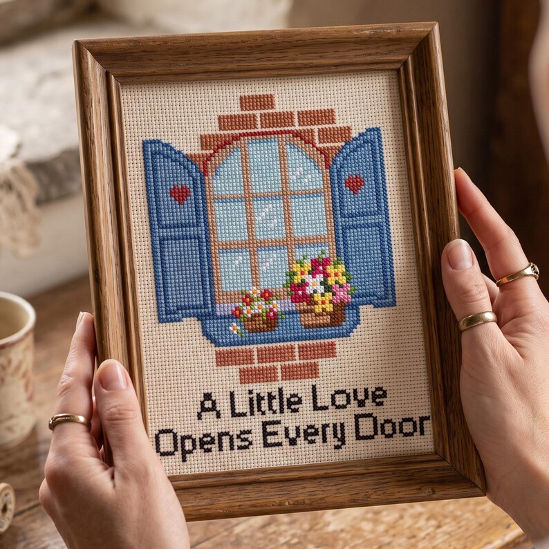 Peut inclure: Une broderie au point de croix encadr&eacute;e repr&eacute;sentant une fen&ecirc;tre avec des volets bleus ouverts, des c&oelig;urs rouges et des jardini&egrave;res sur un mur de briques. Le texte A Little Love Opens Every Door est brod&eacute; en noir sur un fond en tissu cr&egrave;me.