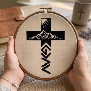 Peut inclure: Un tambour &agrave; broder circulaire en bois contenant un tissu Aida beige avec un motif au point de croix noir. Le motif repr&eacute;sente une croix noire avec une silhouette de soleil et de montagne au sommet et le texte G > N sous la croix.