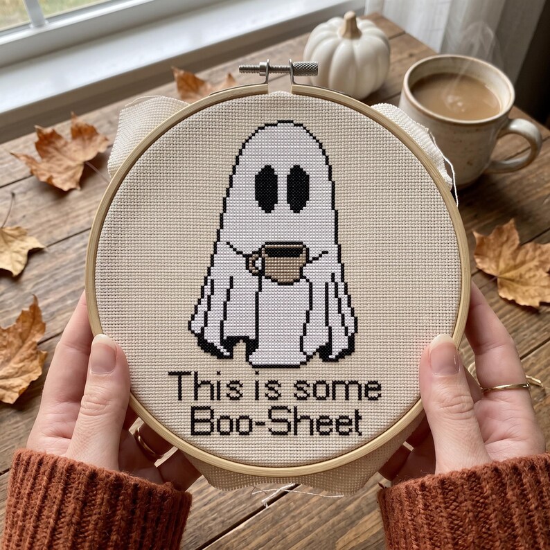 Op de afbeelding: Een ronde houten borduurring met een kruissteekontwerp van een witte geest die een kleine bruine koffiemok vasthoudt. Onder de geest staat de zwarte tekst: This is some Boo-Sheet. De stof is lichtbeige en heeft een duidelijk rasterpatroon.