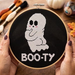 Puede incluir: Un bastidor de bordado circular de madera que contiene un proyecto de punto de cruz sobre tela negra. El dise&ntilde;o presenta un fantasma blanco con ojos negros y mejillas rosadas, sobre el texto blanco pixelado que dice BOO-TY en bloque.