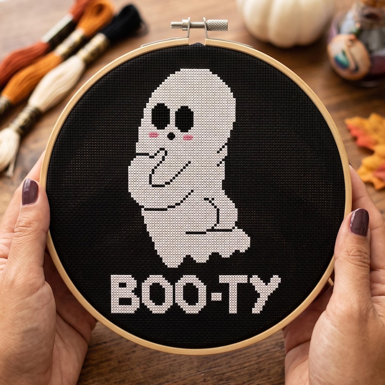 Puede incluir: Un bastidor de bordado circular de madera que contiene un proyecto de punto de cruz sobre tela negra. El dise&ntilde;o presenta un fantasma blanco con ojos negros y mejillas rosadas, sobre el texto blanco pixelado que dice BOO-TY en bloque.