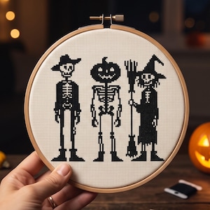 May include: A circular wooden embroidery hoop containing a piece of off-white fabric with a black cross-stitch design. The pattern features three pixelated figures: a skeleton wearing a hat, a skeleton with a pumpkin head, and a witch holding a broom.