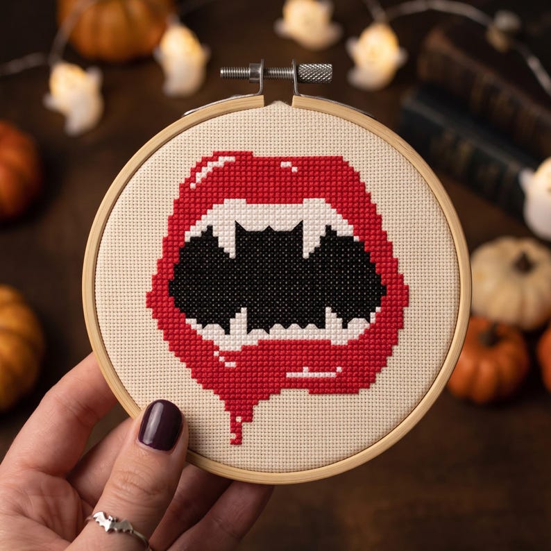 Op de afbeelding: Een kruissteek borduurproject in een ronde houten borduurring met een gepixeld ontwerp van een open mond met rode lippen, scherpe witte hoektanden en een zwarte binnenkant. Het ontwerp is geborduurd op een lichtbeige stoffen ondergrond.