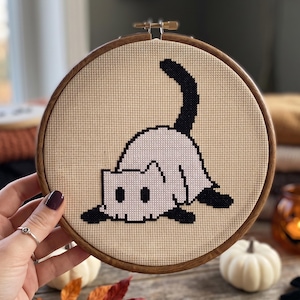 May include: A circular wooden embroidery hoop containing a cross-stitch pattern on beige fabric. The design features a white cat with black ears, paws, and a raised tail, depicted in a pixelated style. The cat is shown in a pouncing position.