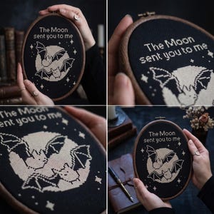 Puede incluir: Un bastidor de bordado ovalado con tela negra y un dise&ntilde;o de punto de cruz de dos murci&eacute;lagos frente a una luna grande. El texto "The Moon sent you to me" est&aacute; bordado en blanco sobre la luna, rodeado de peque&ntilde;as estrellas blancas.