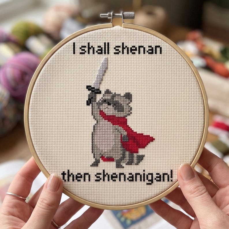Pu&ograve; includere: Un telaio da ricamo circolare in legno contenente un disegno a punto croce su tessuto color crema. Il disegno raffigura un procione grigio con un mantello rosso che impugna una spada grigia, con il testo nero I shall shenan then shenanigan!.