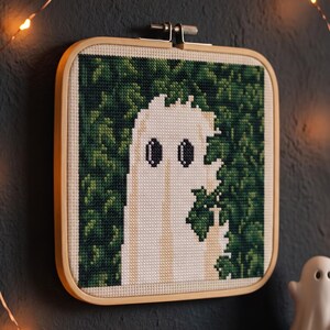 May include: A square cross-stitch embroidery piece in a light wood frame featuring a white ghost figure with dark oval eyes set against a dense, dark green leafy background. The ghost is depicted with a jagged, pixelated edge and a raised hand gesture.