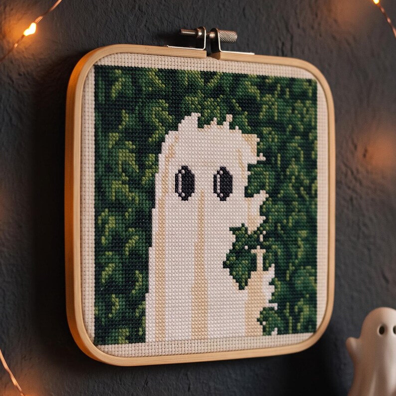 May include: A square cross-stitch embroidery piece in a light wood frame featuring a white ghost figure with dark oval eyes set against a dense, dark green leafy background. The ghost is depicted with a jagged, pixelated edge and a raised hand gesture.